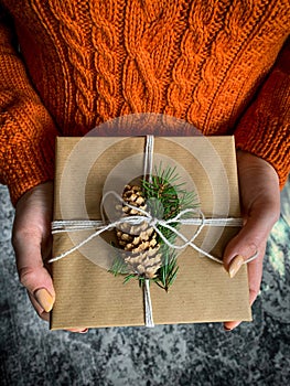 Female`s hands in pullover holding Christmas gift box decorated with evergreen branch and pine cone.