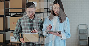 A female sales manager using a tablet computer is talking to a male worker holding a cardboard package. A warehouse with