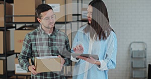A female sales manager using a tablet computer is talking to a male worker holding a cardboard package. A warehouse with