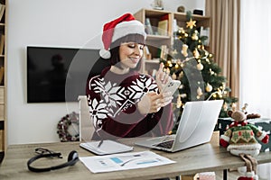 Female in Santa hat holding smartphone while staying in workplace at home.