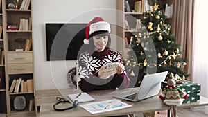 Female in Santa hat holding smartphone while staying in workplace at home.