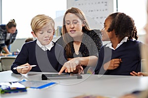 Female school teacher helping two kids using a tablet computer at desk in a primary school classroom, front view