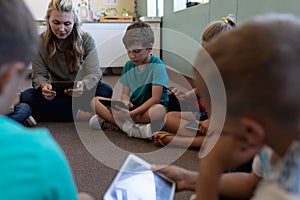 Female school teacher sitting cross legged on the floor in a circle with a group of schoolchildren a