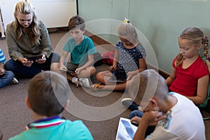 Female school teacher sitting cross legged on the floor in a circle with a group of schoolchildren a