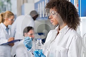 Female Scientific Researcher In Laboratory, African American Woman Working With Flask Over Group Of Scientist Making