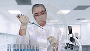 A female scientist examines a blue fluid sample using a micropipette and test tubes while sitting at a table in the