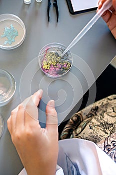 Female scientist hands adding drop of liquid from from pipette over petri dish with glitters mix