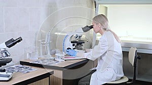 a female scientist in the laboratory conducts a study looking through a microscope recording readings in a journal.