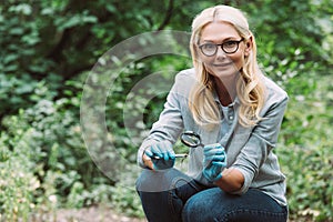 female scientist in latex gloves sitting with twig in tweezers and magnifier