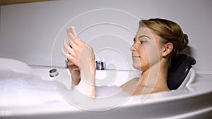 Female scrolling social media, lying in bathtub with foam bubbles, relaxation