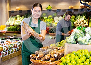Female seller holding yuca standing in fruit and vegetable section of supermarket