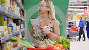 Female shopper with a trolley in the aisle of a store with a shopping list while shopping for food. Woman reading in her