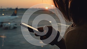 Female silhouette using smartphone. Close up shot of woman hands with mobile at sunset or sunrise background at airport