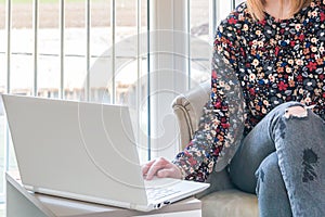 Female sitting in armchair and typing on white keyboard. Woman working at home office and using lap top or notebook.