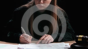Female sitting at the desk with wooden hammer and paper documents, dark room bright spotlight. Woman judge in the court
