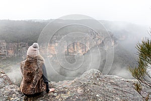 Female sitting on mountain top cliff ledge looking out into the misty fog