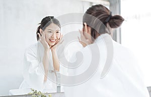 Female Skin Care. Young asian woman touching her face and looking to mirror in bathroom.