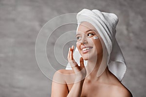 Female skincare concept. Lady applying moisturizer cream under eyes, standing in bathroom, looking aside and smiling