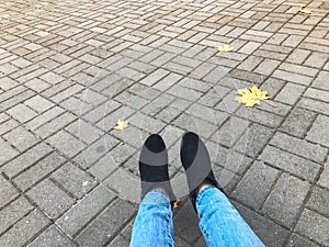 Female slender legs in jeans and boots, shoes against a background of gray stone concrete paving slabs and autumn yellow leaves.