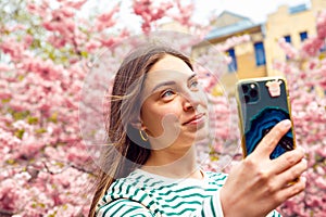 Female with smartphone taking selfie in cherry flower blossom garden. Young smiling european white caucasian woman with mobile