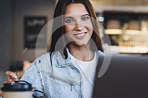 Female smiling, looking at the camera. Attractive business woman sits at table in front of laptop talks on mobile phone