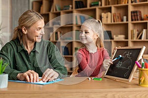 Female speech therapist teaching happy little girl alphabet, child writing word cat on blackboard with chalk