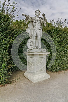 Female Statue in beautiful garden. Monument made of white stone stands on rectangular pedestal. Statue of woman which holds out