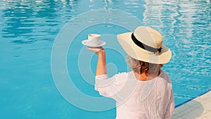 female in a straw hat holds a cup of coffee in her hand over a blue swimming pool concept of good morning and day