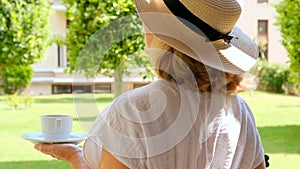 female in a straw hat holds a cup of hot drink and drinks coffee, sitting in the morning on a summer terrace on a sunny