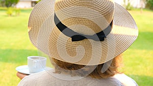 female in a straw hat holds a cup of hot drink and drinks coffee, sitting in the morning on a summer terrace on a sunny