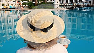 female in a straw hat wearing a white dress relaxing by the pool with a cup of coffee, vacation concept, good morning