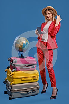 Female in straw hat, white blouse, red pantsuit, black heels. Holding passport and ticket, posing on blue background