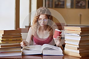 Female student posing with coffee in hand, suraunded with stacks of books at reding room. Young woman sitting at table doing