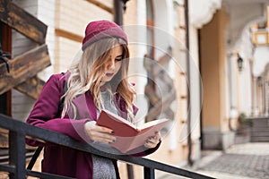 A female student in a purple coat concentrates reading a textbook on the street