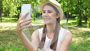 Female student sitting on grass on college campus having video chat via online app on mobile phone