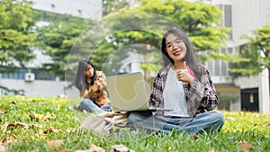 A female student is sitting on the grass, showing her thumb up, and using a laptop for studying