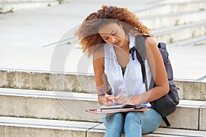 Female student sitting outside on steps with bag and writing in book