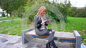 Female student is sitting on parkbench and e-learning using her tablet PC. She is working with web app or passing the