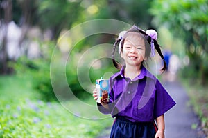 Female student standing holding blue milk carton box. Sweet smiley girl wearing a purple school uniform.