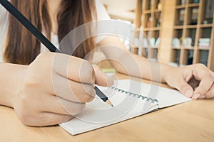 Female student taking notes from a book at library. Young asian woman sitting at table doing assignments in college library.