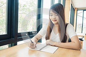 Female student taking notes from a book at library. Young asian woman sitting at table doing assignments in college library