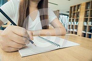 Female student taking notes from a book at library. Young asian woman sitting at table doing assignments in college library.