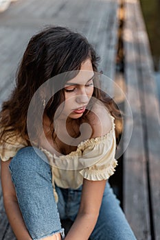 Female summer portrait of a beautiful stylish woman with curly hair in fashionable blue jeans and yellow top sits on a wooden pier