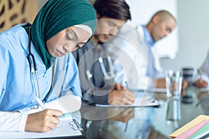 Female surgeon in hijab writing on clipboard at table in the hospital