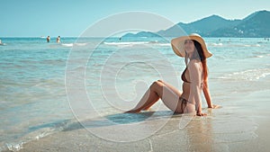 Female in swimsuit and hat sits on shore in water, enjoying seascape tropical island. Feet immersed in azure water