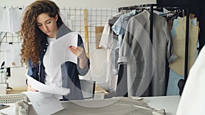 Female tailor is rolling out fabric bolt on studio table then taking garment sketch and drawing on paper. Designing