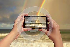 Female taking picture on mobile phone of double rainbow over ocean and tropical beach