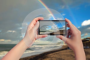 Female taking picture on mobile phone of double rainbow over ocean and tropical beach