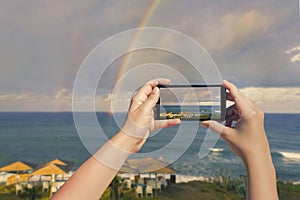 Female taking picture on mobile phone of double rainbow over ocean and tropical beach with umbrellas chairs and tables