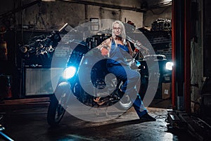 Female tattooed mechanic in work overalls hold a big wrench and posing for a camera in garage